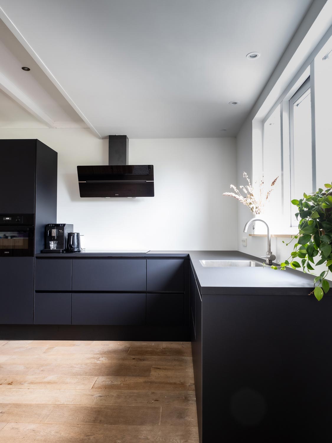 An image of a modern kitchen design with dark, handleless cabinetry in an L-shape. Image features an extractor hood over an integrated induction hob, an integrated oven and a Quooker tap.