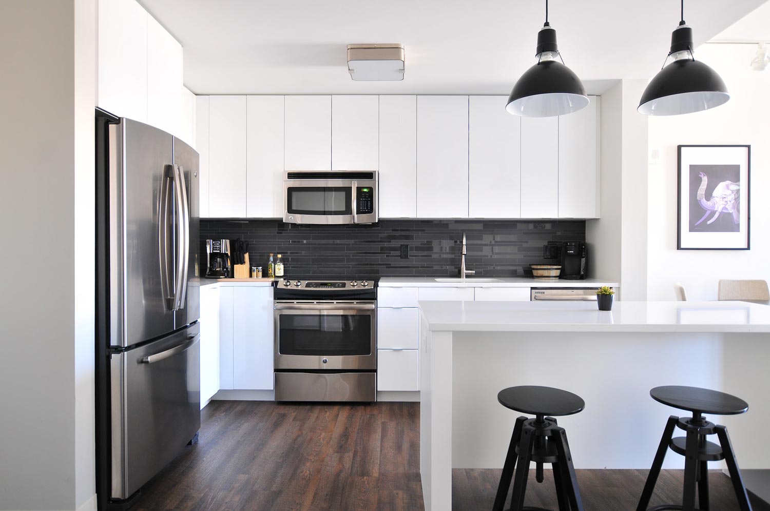 An example of a modern kitchen design, with white cabinetry, integrated microwave, oven and dishwasher and freestanding fridge/freezer. To the right, a kitchen island with two dark wood stools