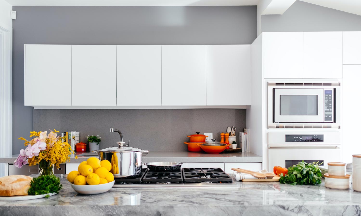 A modern kitchen design with grey walls and white cabinetry. In the foreground is a kitchen island with a built-in gas hob, surrounded by decorative food items. 